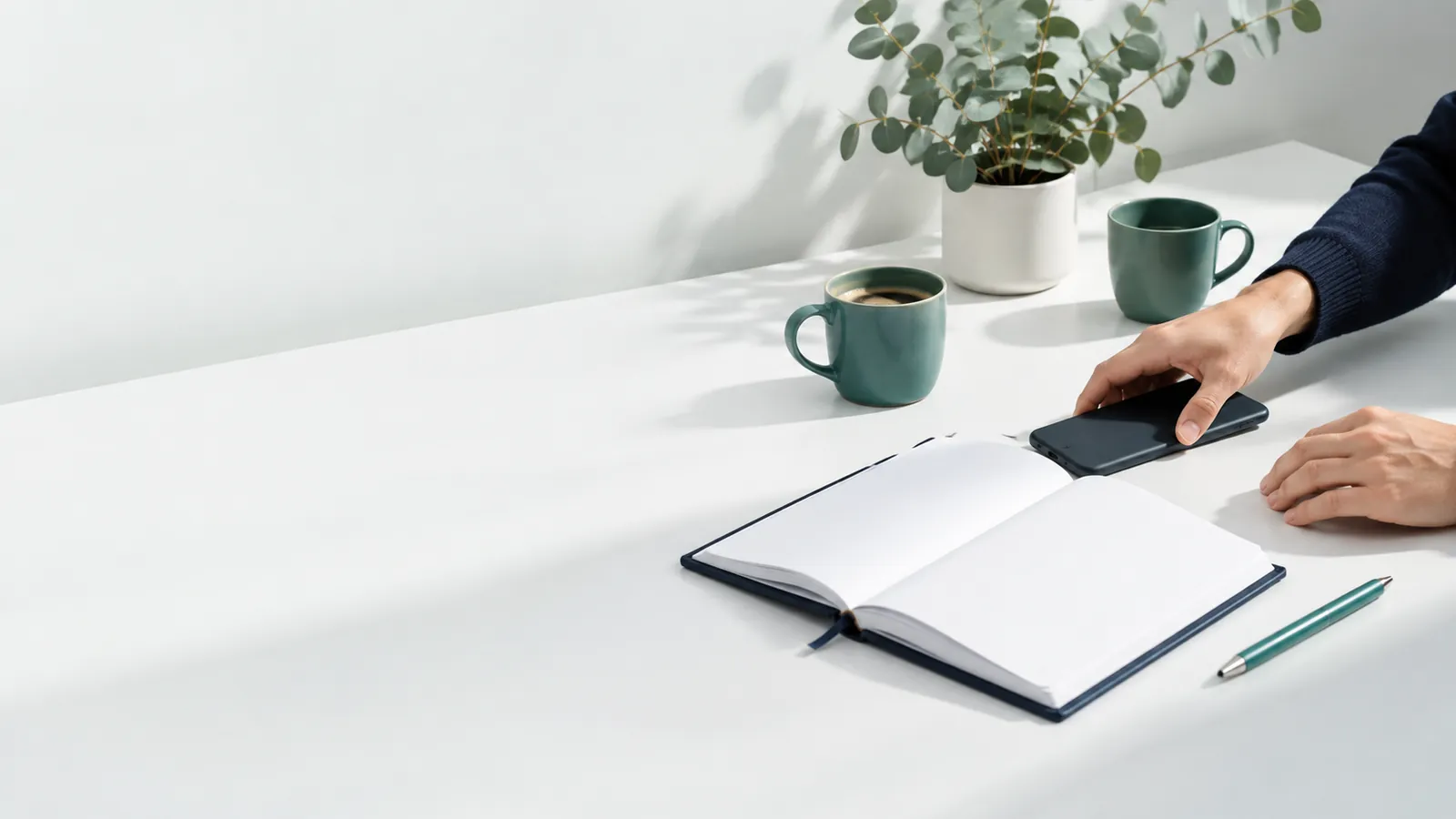 Les mains d'une community manager posent un smartphone, écran retourné, à côté d'un carnet de planification et de tasses vertes, dans un studio clair