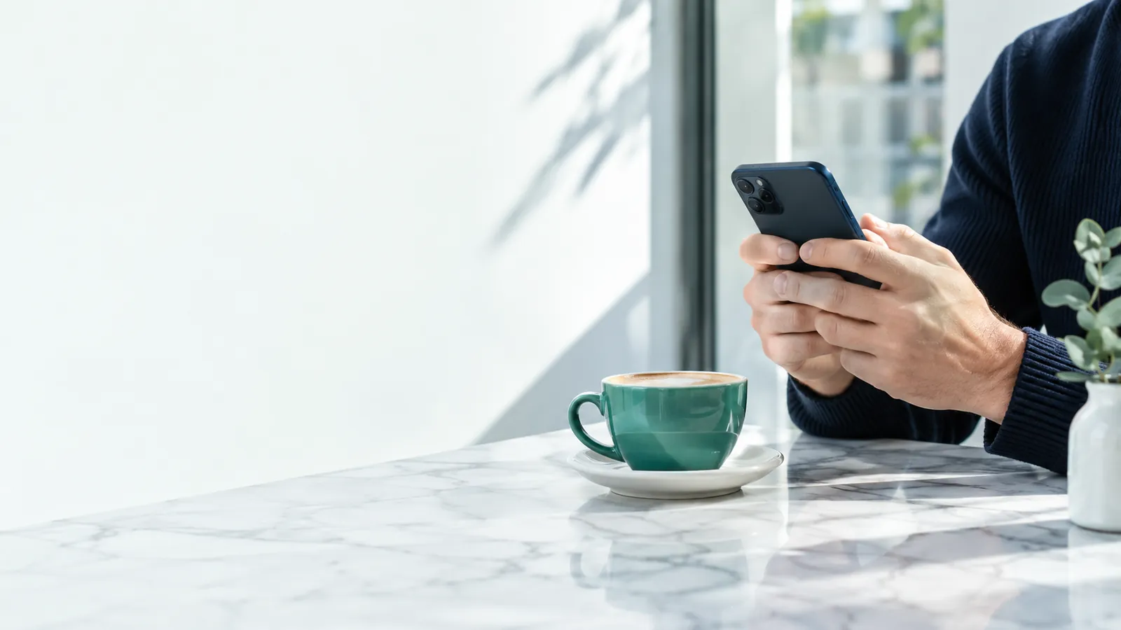 Les mains d'une personne tiennent un smartphone, écran tourné vers elle, à une table de café claire près d'une tasse verte