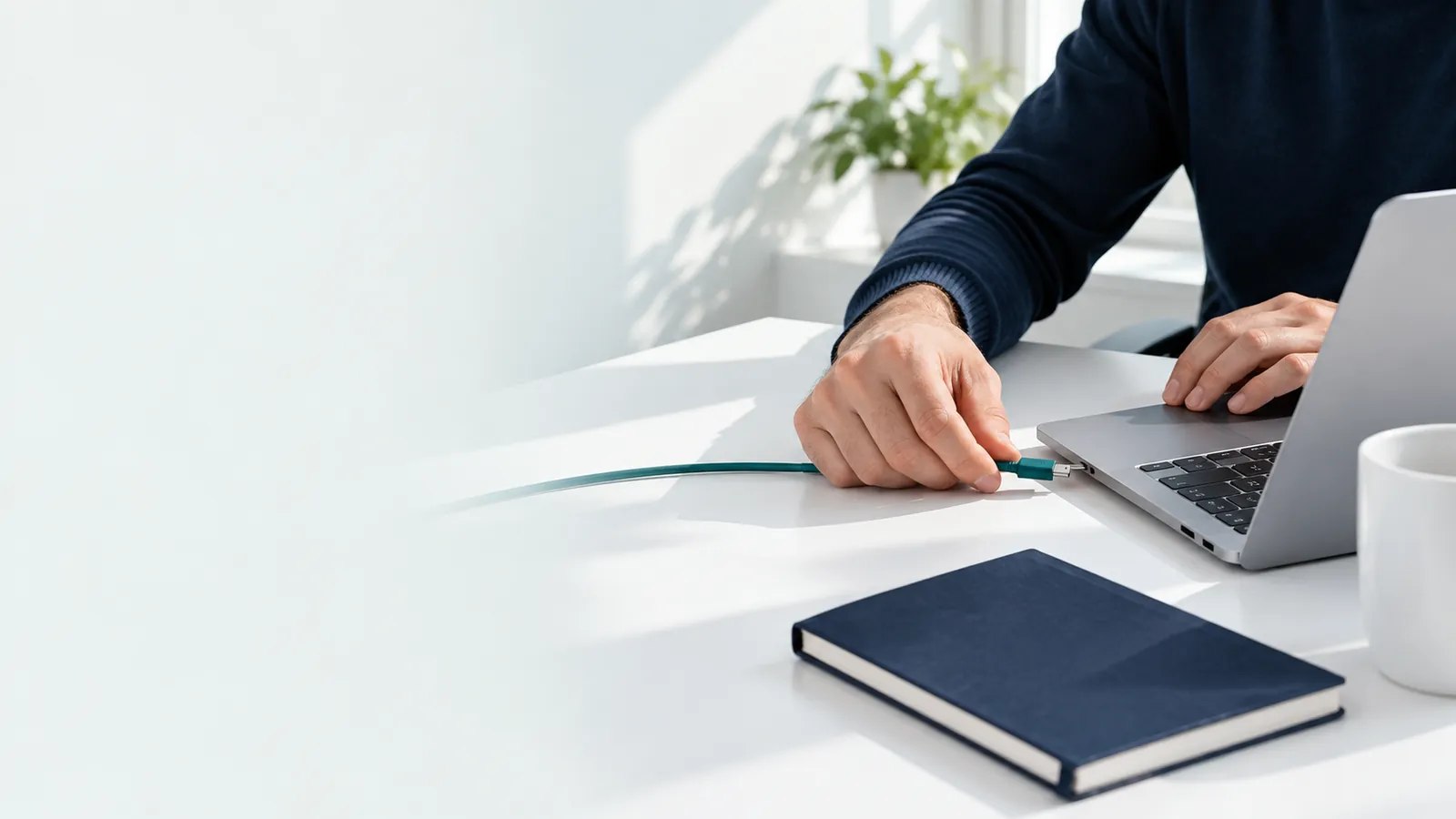 Les mains d'un gérant branchent un câble vert sur le côté d'un ordinateur portable, écran tourné vers l'arrière, sur un bureau blanc
